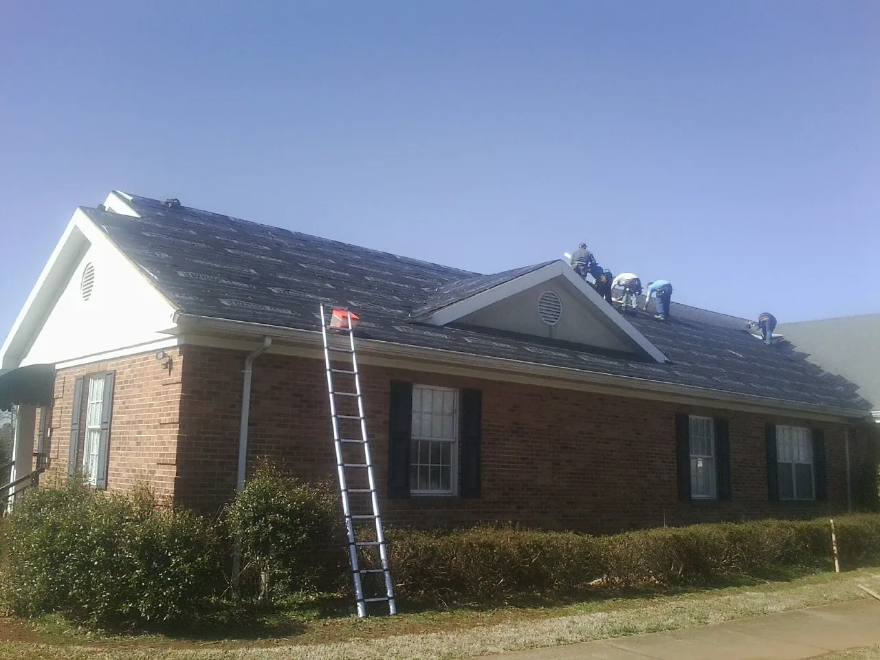 Crew tearing shingles off a brick ranch roof with a ladder set — Emergency Roof Repair work by our crew in Drums