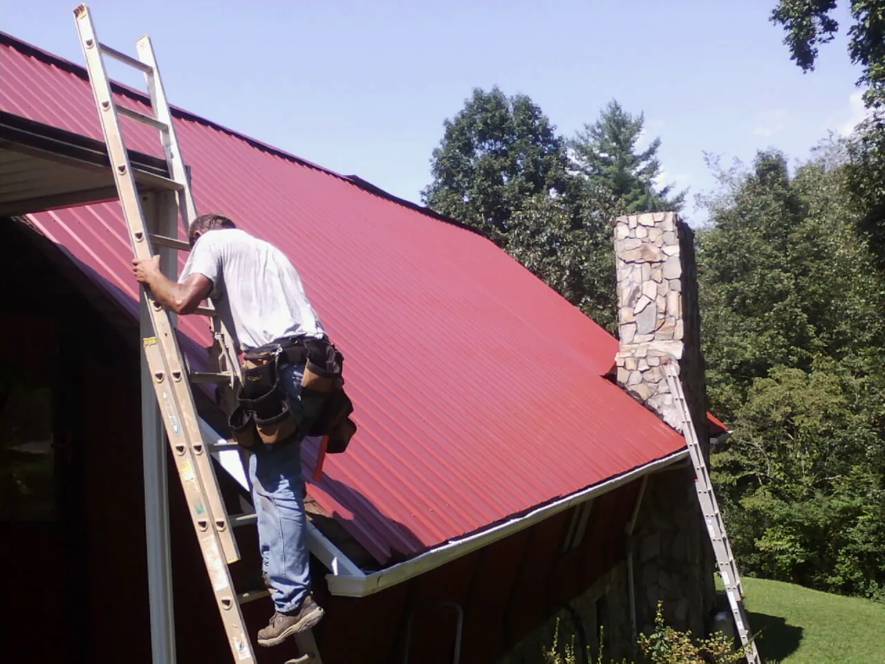 Roofer on a ladder beside a red metal roof and stone chimney — Soffit & Fascia Repair by Roofing Brothers in Drums